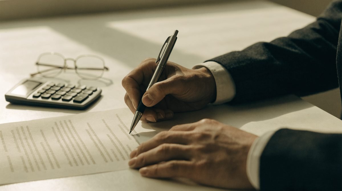 Mãos segurando caneta sobre contrato impresso, com calculadora e óculos sobre a mesa